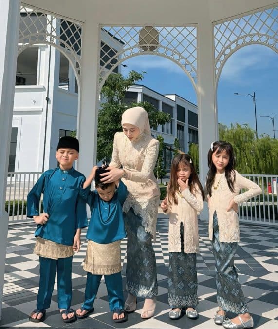 Mother in a Beige Lace Outfit and Hijab Helps Two Young Boys in Blue Baju Melayu While Two Girls in Cream Kebaya Smile Nearby on a Black and white Checkered Patio Under White Decorative Arches