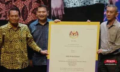Three men stand on stage holding a large framed certificate with a crest at the top, posing for a photo.