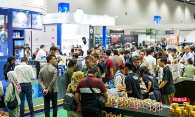 Crowded trade show hall with multiple booths and attendees sampling beverages at a busy stand.