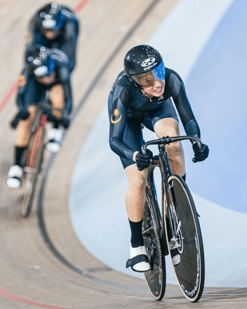 Female track cyclist in a black skinsuit and helmet leans into a bend on an indoor velodrome, with trailing riders in the background.