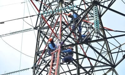 Crew of workers in hard hats climbing a metal transmission tower with insulators and cables.