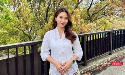Woman in a white blouse posing outdoors beside a black railing with colorful trees behind her.