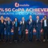 Seven professionals in suits wave at the camera on a stage with a blue digital backdrop reading '90% 5G CoPA ACHIEVED 9 MONTHS AHEAD OF SCHEDULE' and a mobile logo, celebrating a milestone.